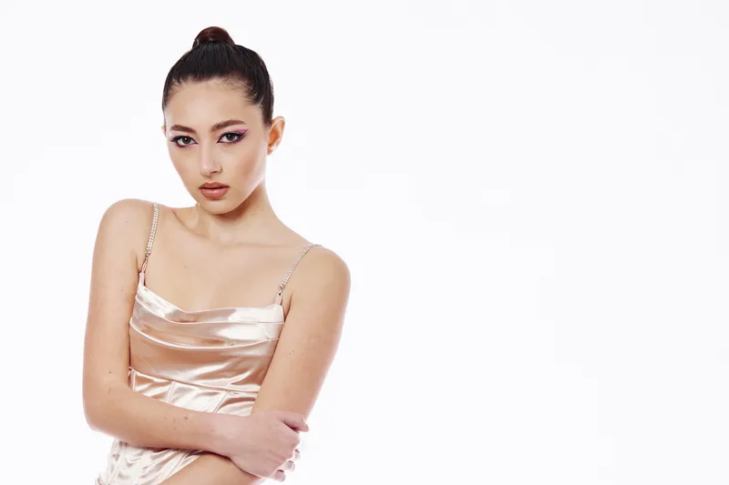 Young woman with silk champagne dress poses thoughtfully in studio. Makeup with silvery eyeshadows, hair sleeked back in a high bun. Creative beauty fashion concept.