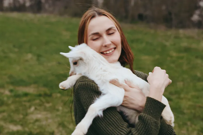 A smiling young woman embraces a white goat kid outdoors in a grassy field, capturing a warm, candid moment of rural lifestyle, affection, and animal companionship.