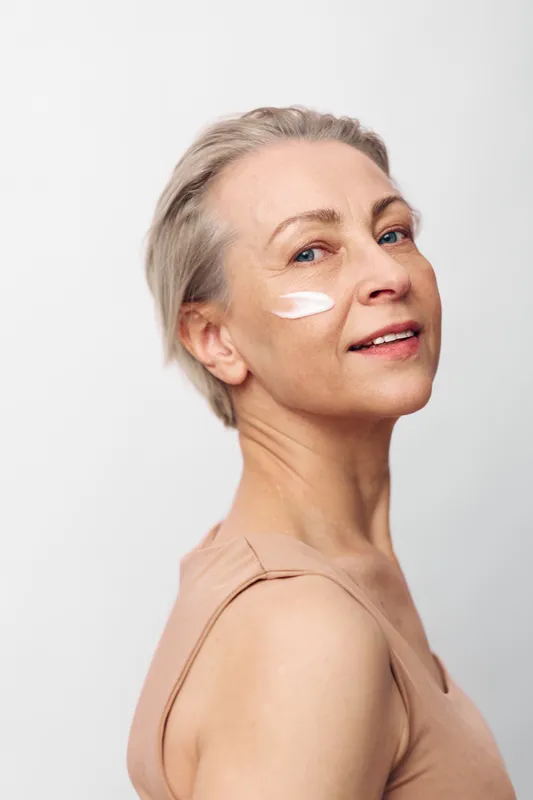 Mature woman with short grey hair applies white cream on cheek, smiling gently. Clean skin and natural beauty concept with minimal makeup on light background for skincare and wellness.