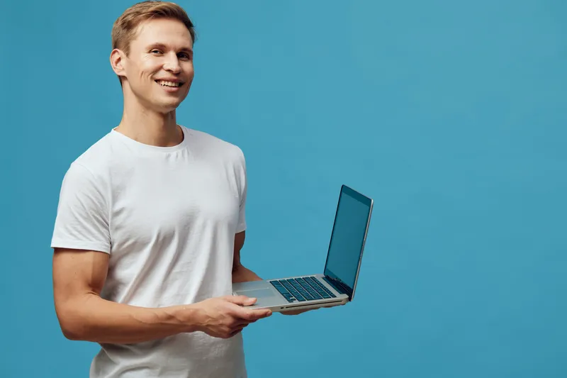 Smiling young man holding a laptop computer and looking at the camera isolated on plain blue background. Casual clothing, positive expression, technology, happiness, modern lifestyle, workspace concep