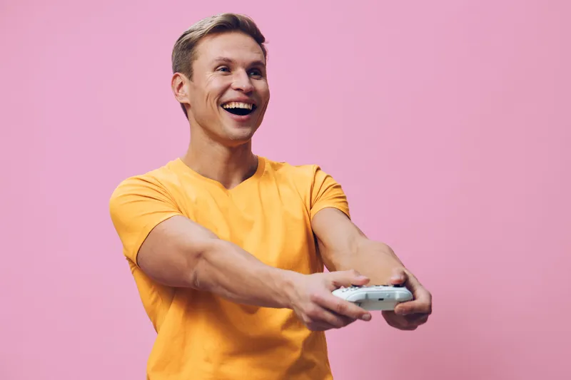 Smiling young man with short blond hair wearing bright yellow t-shirt holding game controller, expressing joy and excitement, isolated on pink background, studio portrait of male gamer, lifestyle conc