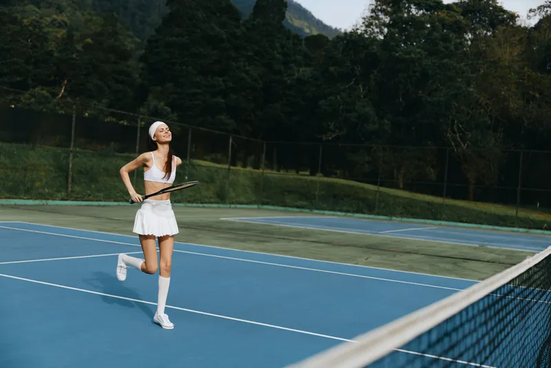 Young female tennis player in white sportswear joyfully running on a blue court, showcasing athleticism and energy against a lush green background