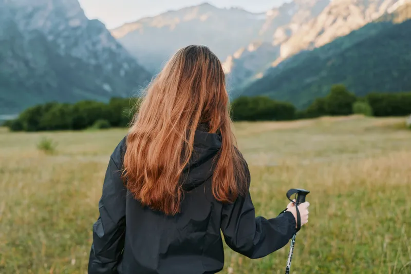 A hiker with long hair wearing a dark jacket stands in a meadow, facing distant mountains under a clear sky, ready to explore and enjoy the outdoors.