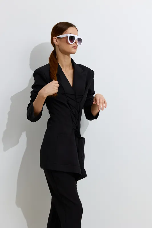 Fashion woman wearing black blazer and pants with white sunglasses posing confidently against white background in stylish minimalistic look.