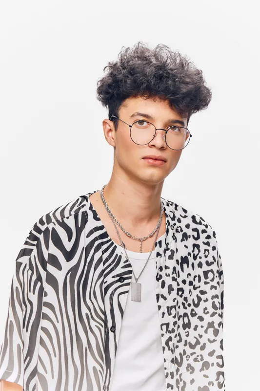 young man with curly hair wearing round glasses and a black and white animal print shirt standing against a white background with a neutral expression