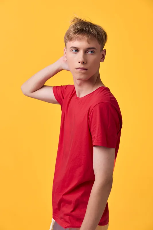 Young man in a red t shirt stands against a bright yellow background, casual pose suggesting contemplation and curiosity, studio shot with vibrant color contrast and minimal styling