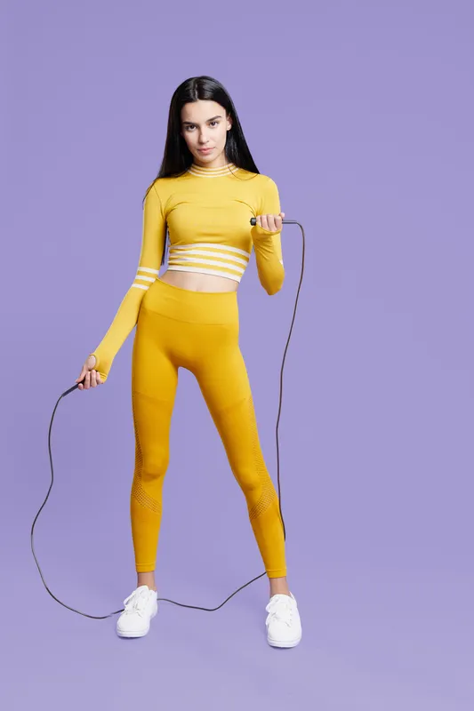Woman sport fitness wearing yellow activewear with long sleeves and leggings, holding a jump rope, standing against plain purple background with focused expression.