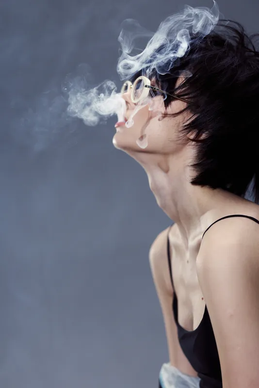 Young woman with short hair blowing smoke against grey background, wearing glasses and a black top, conveying mystery and edginess.