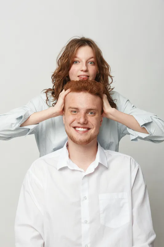 Smiling young man with red hair sits in front of woman with curly hair holding his head. Both wear white shirts on light background. Concept of friendship, fun, and casual portrait.