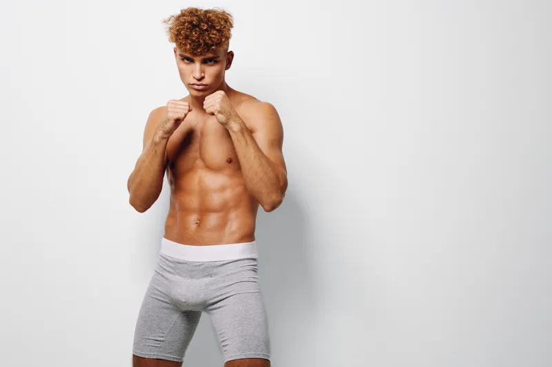 Young athletic man with muscular body posing in fighting stance wearing gray shorts against white background. Fitness, strength and boxing training concept with confident expression.