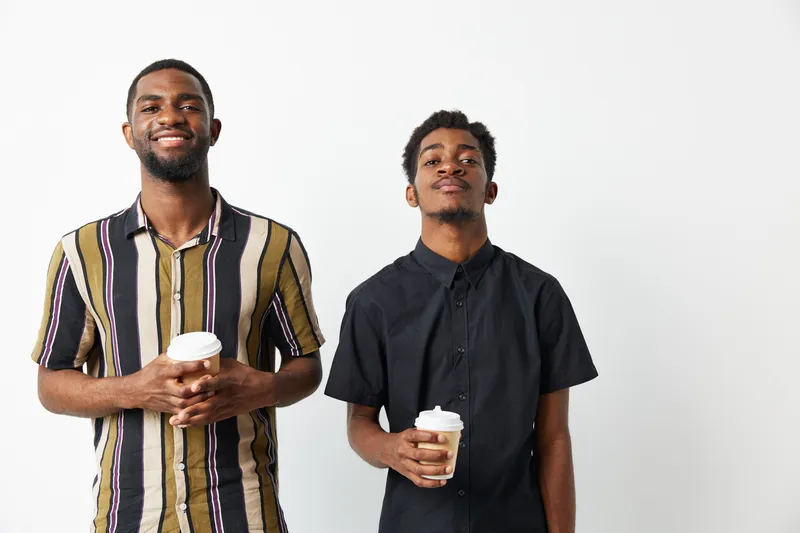 Happy African American men standing together, holding coffee cups and smiling. Dressed casually in stylish shirts, they project a fun and relaxed vibe.
