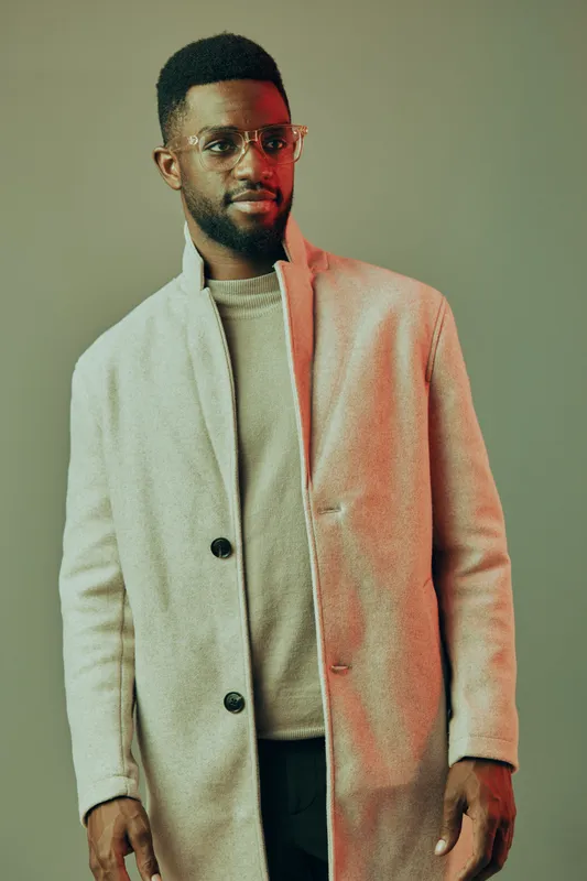 Stylish African American man in modern coat and glasses posing against soft gradient background, showcasing contemporary fashion and confident expression