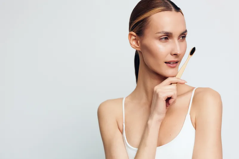 Young woman with clear skin holding facial brush near face, natural makeup, white tank top, looking aside, minimalistic background, beauty skincare concept, soft lighting