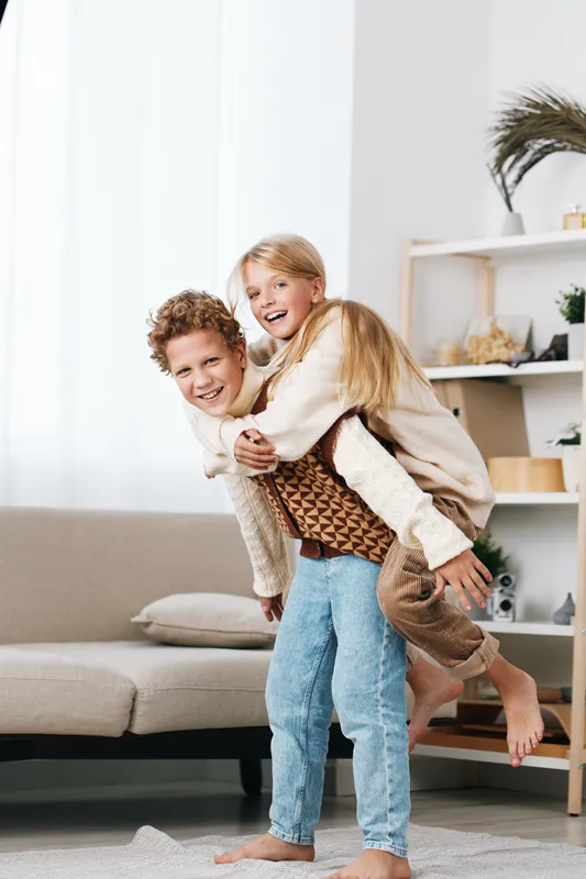 Playful siblings share a joyful piggyback moment in a bright living room, capturing candid smiles, warm textures, and a carefree, family vibe in a cozy home setting.