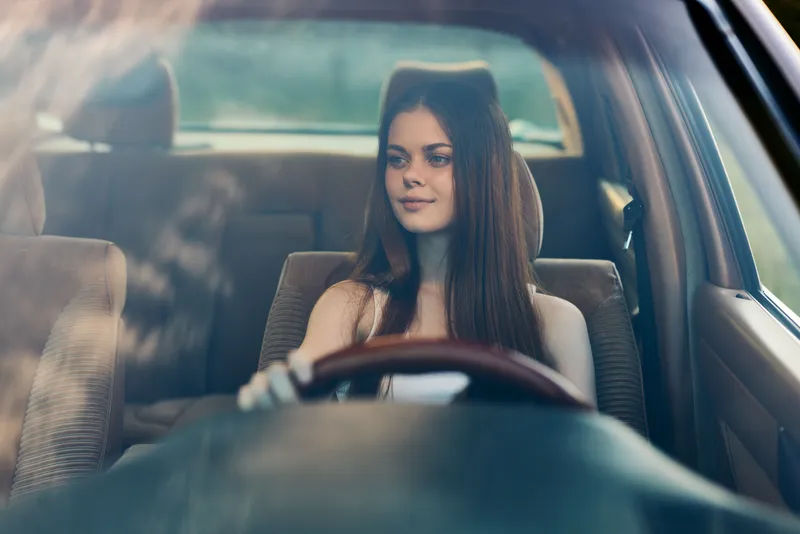 Young woman sitting in a car, smiling while holding the steering wheel, reflecting a sense of freedom and adventure on a sunny day.
