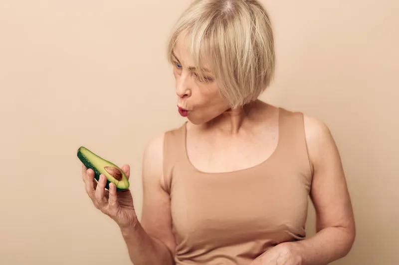 Mature woman with short blonde hair examines fresh avocado half with curiosity against beige background. Concept for healthy lifestyle, nutrition, and natural food choices in middle age.