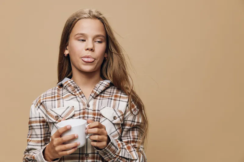 Young woman with long hair wearing plaid shirt holds white cup with both hands while making playful face against beige background.