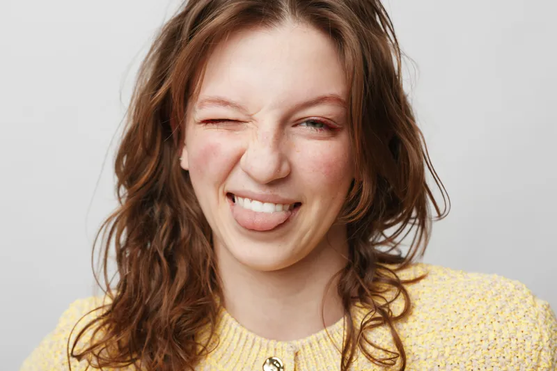 Young woman with wavy brown hair wearing yellow sweater makes playful face sticking out tongue and winking. Studio portrait on light background with casual and fun mood.