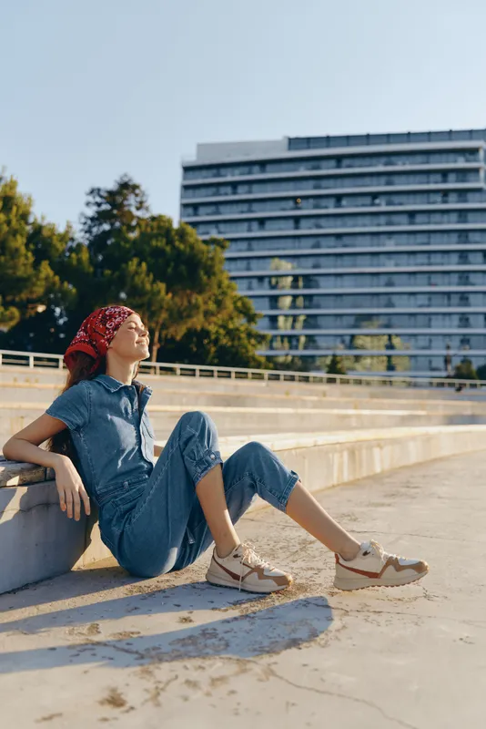 Lifestyle woman in modern boho-western denim street style relaxes sunbathing outdoors with a vibrant red bandana and casual shoes in film color tones.