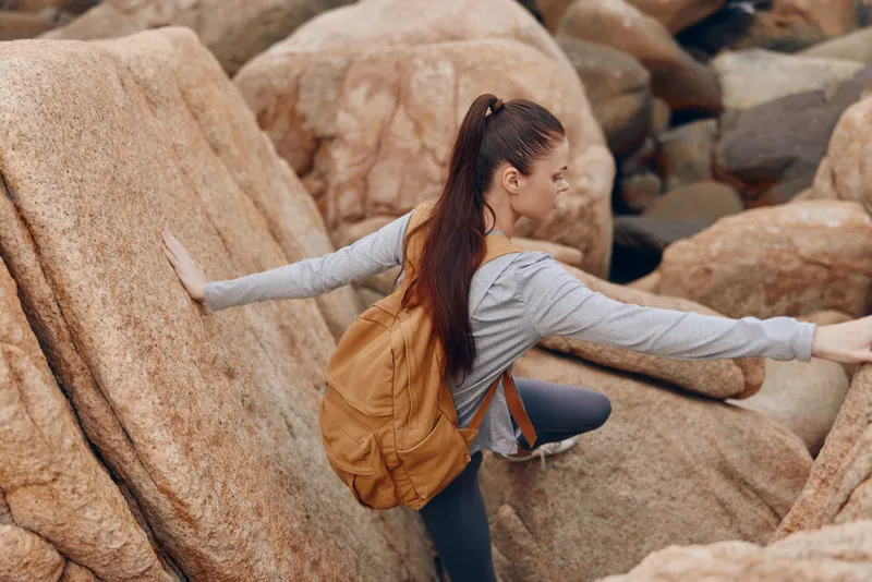 Adventurous young woman climbing rocky terrain in outdoor fashion with a backpack, showcasing determination and joy amid natural scenery