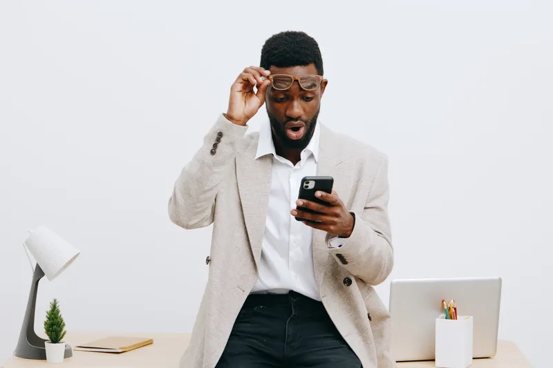 Professional young African-American man in casual business attire looking surprised while using smartphone in modern office environment with minimal decor, white background. Business lifestyle concept