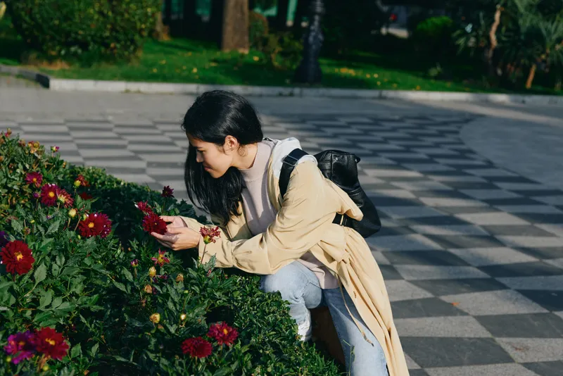 A female gardener kneels beside flowers in a park, selecting petals with focus and care, showcasing gardening, outdoor activity, and nature appreciation in a calm daylight setting