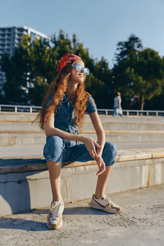 Lifestyle woman in modern boho-western denim street style sits confidently outdoors wearing sunglasses and a red bandana in natural film color tones.
