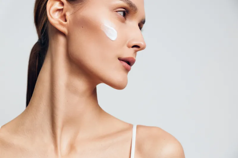 Profile of a young woman with clear skin applying white face cream on cheek. Minimalist background with soft lighting emphasizes skincare and natural beauty concept.