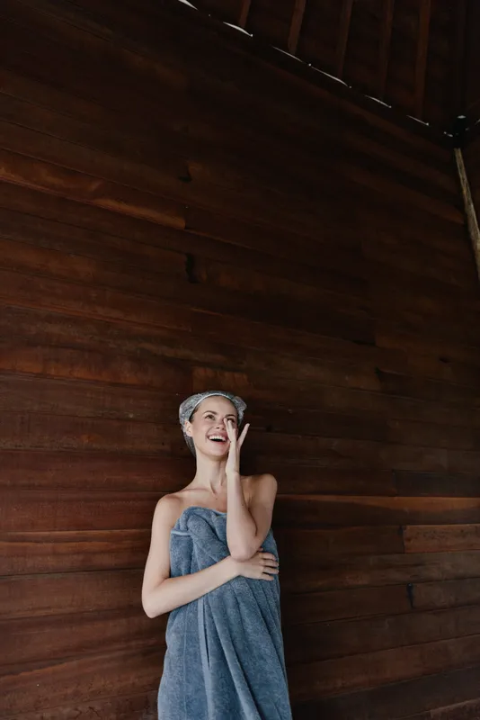 smiling woman wrapped in towel with headband stands against dark wooden wall, expressing joy and relaxation indoors in a cozy atmosphere