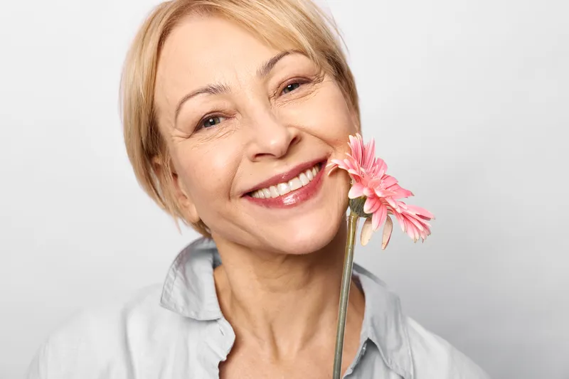 Woman with short blond hair smiling in closeup portrait, holding a pink flower by her cheek, beauty spring mood, soft studio light, wellness