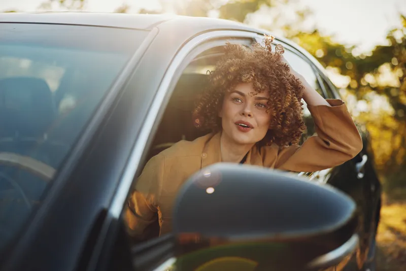 Smiling woman with curly hair leaning out of a car window during golden hour, enjoying a carefree moment. Natural light and warm tones convey lifestyle, travel, and self expression.