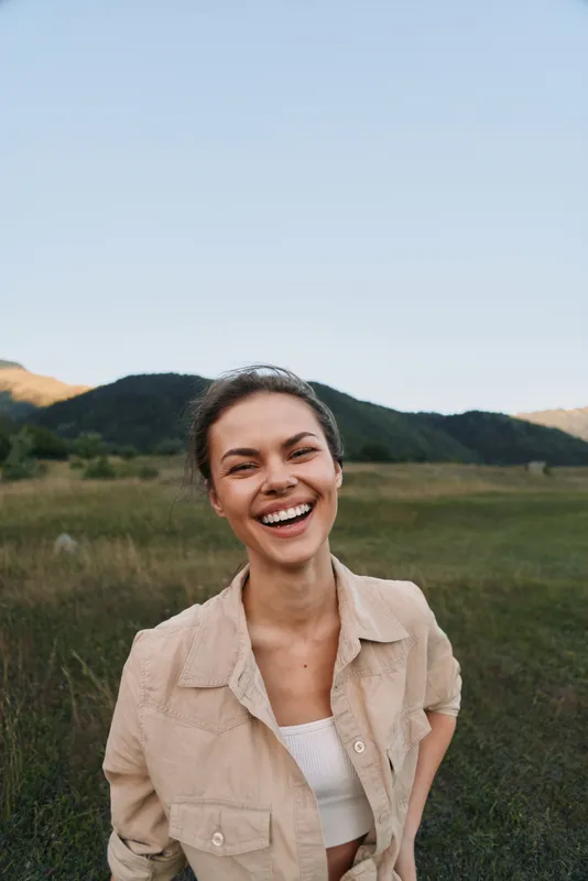 Happy woman outdoors smiling broadly with relaxed posture in natural landscape during daytime, casual outfit, clear sky, green hills background