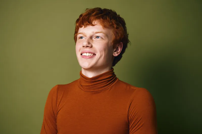 Young man with red hair wearing brown turtleneck smiling and looking aside with green background. Portrait of confident casual male with natural expression in studio light for positive mood.