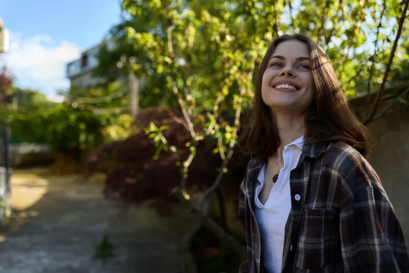 Young woman with long dark hair smiles up at the sunny sky as she stands in a leafy outdoor setting wearing a plaid shirt over a white tee, radiating relaxed carefree mood