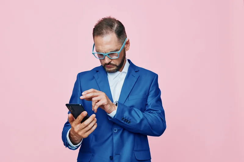 A professional man in a blue suit stands against a solid pink background, focused on his smartphone, conveying concentration and determination in a modern business setting.