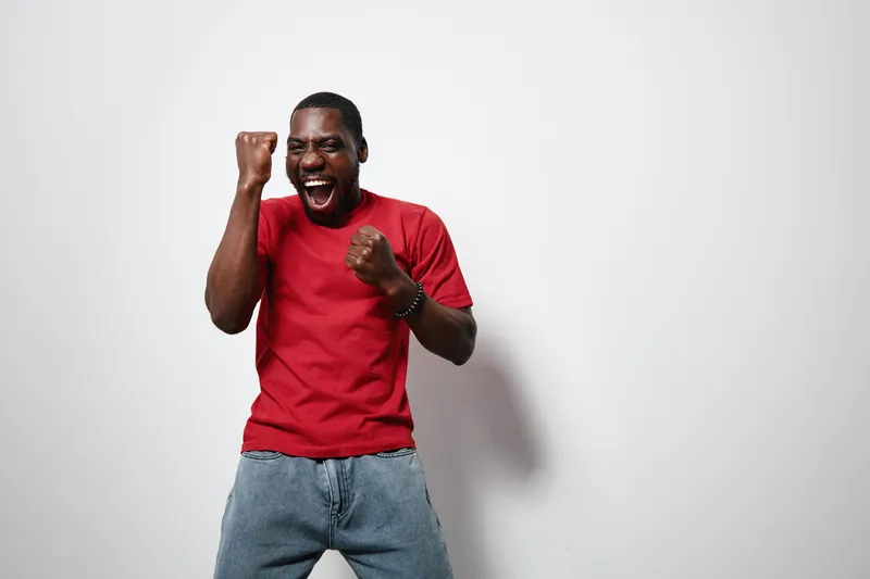 African American man with dark skin wearing red t shirt and jeans celebrating with fists clenched and eyes closed on white background. Excited, happy, casual, isolated studio portrait.