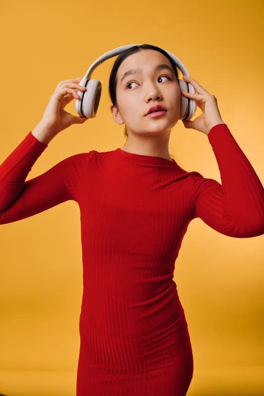 Young woman wearing red dress and white headphones stands against orange background, adjusting audio device with thoughtful expression. Casual outfit, technology usage and music listening concept.