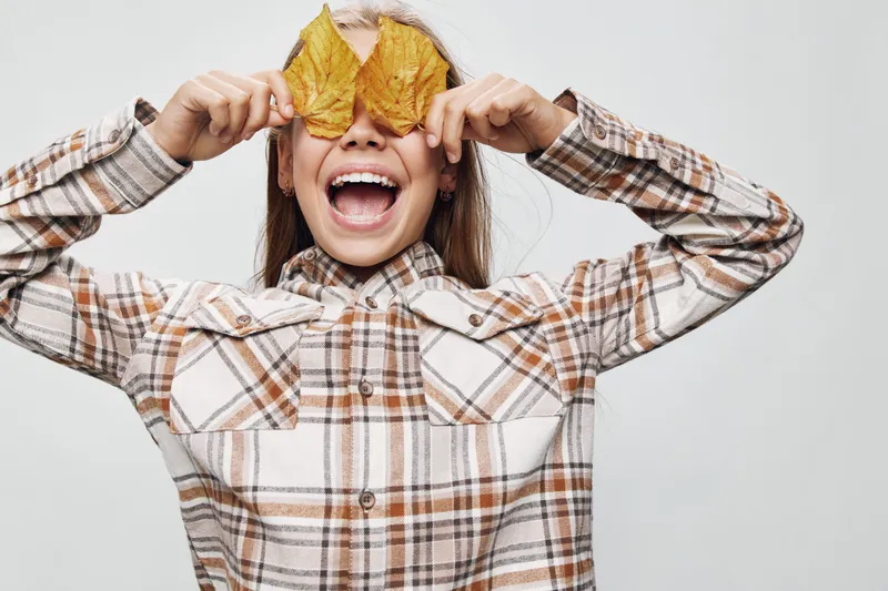 Girl wearing plaid shirt holding autumn leaves over eyes smiling with open mouth isolated on white background. Child playing with fall foliage in casual clothes for seasonal theme.