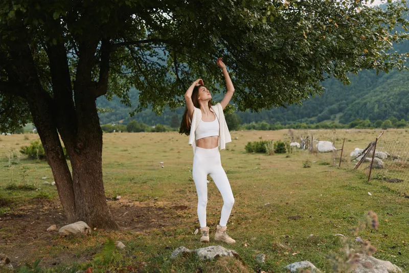 Young woman in white clothes stretches arms and enjoys nature standing near tree in green meadow with mountains in background during daytime outdoors.