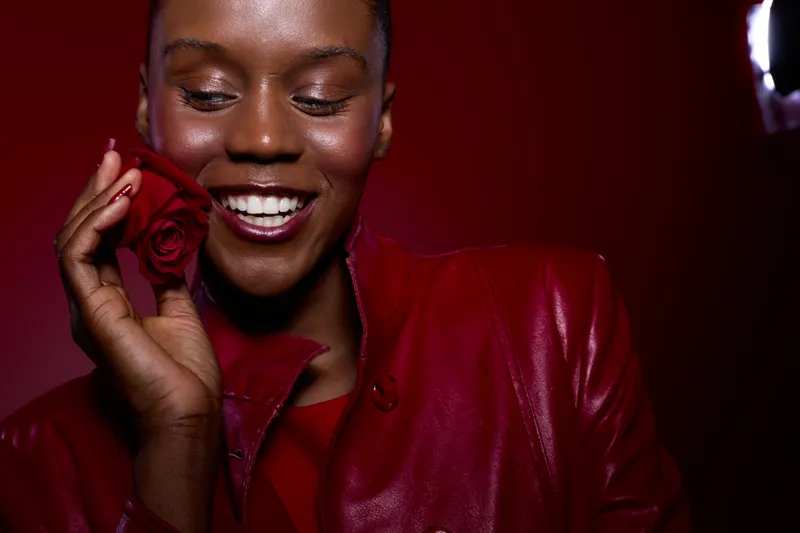 Smiling dark skinned African woman holds red rose near face wearing red leather jacket. Studio portrait on red background. Concept for beauty, happiness, love, and fashion.