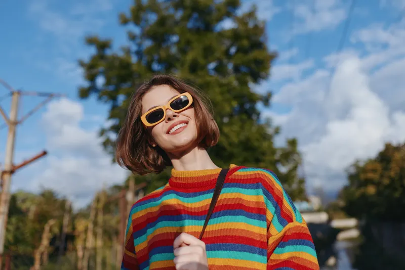Woman portrait with sunglasses and colorful sweater smiling outdoors against blue sky. Young happy fashion look, casual style and sunny mood in park setting.