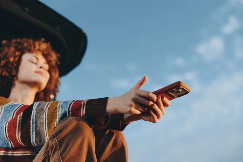 Woman lifestyle outdoors, wearing a hat and sweater, holding a smartphone, looking up toward blue sky, sunlight in soft clouds, casual communication, travel moment, relaxed mood today