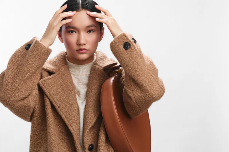 asian woman is seen in a fashion portrait, wearing a warm coat and bag, adjusting her hair with a calm gaze, presenting a modern lifestyle style in studio lighting.