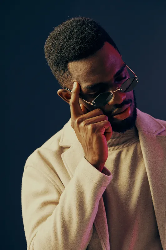 Stylish young Black man in a light coat and glasses, contemplating with a thoughtful expression against a dark background Modern fashion concept