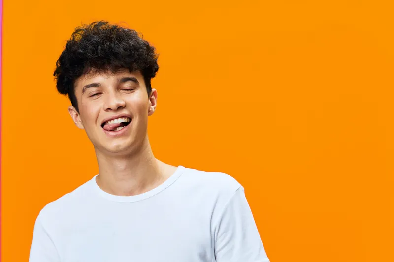 Young man with curly hair making a playful face and sticking out his tongue against a bright orange background, wearing a white t-shirt, joyful expression.