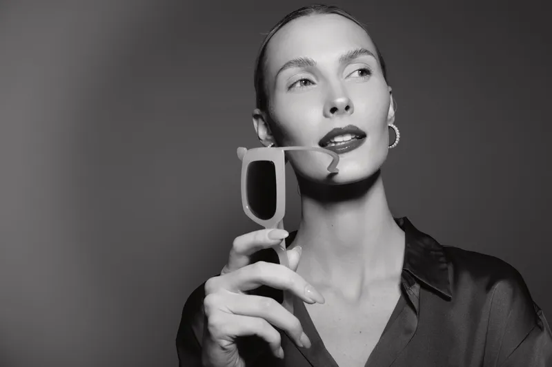 Elegant white woman with pale skin holding sunglasses near face, wearing dark shirt and hoop earrings in black and white portrait.