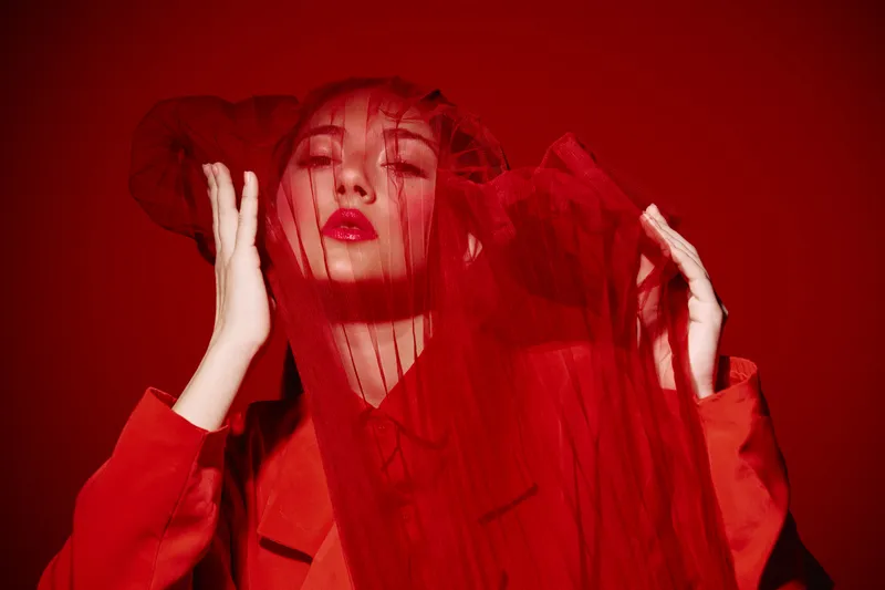Elegant woman wearing vibrant red coat and sheer red veil posed with intense gaze in bold creative fashion portrait with monochromatic crimson background.