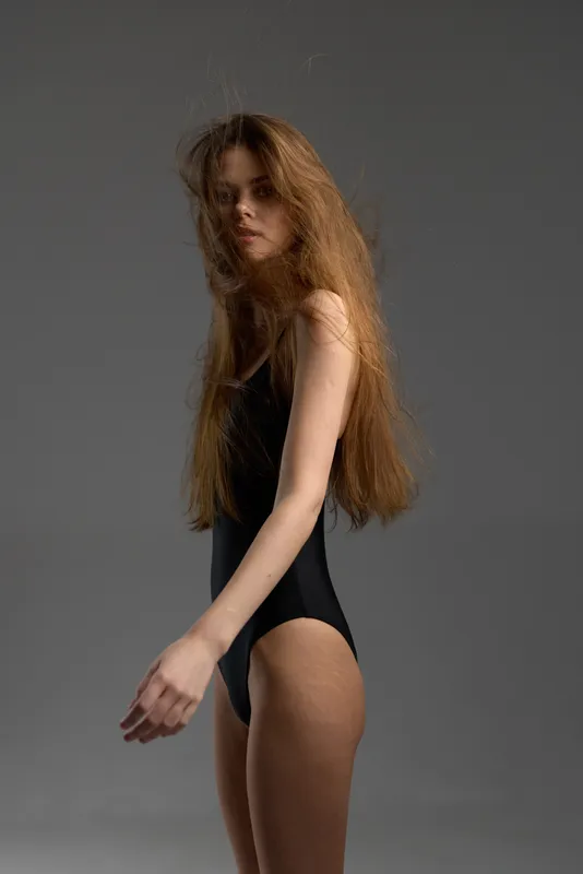 Young woman with long hair in black bodysuit posing with serious expression against grey background