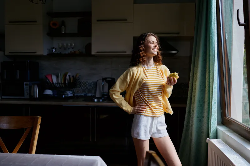 Cheerful woman indoors in a sunlit kitchen, standing by a window with a relaxed pose, wearing casualwear, enjoying a moment of home lifestyle and natural warmth, candid attention on everyday life