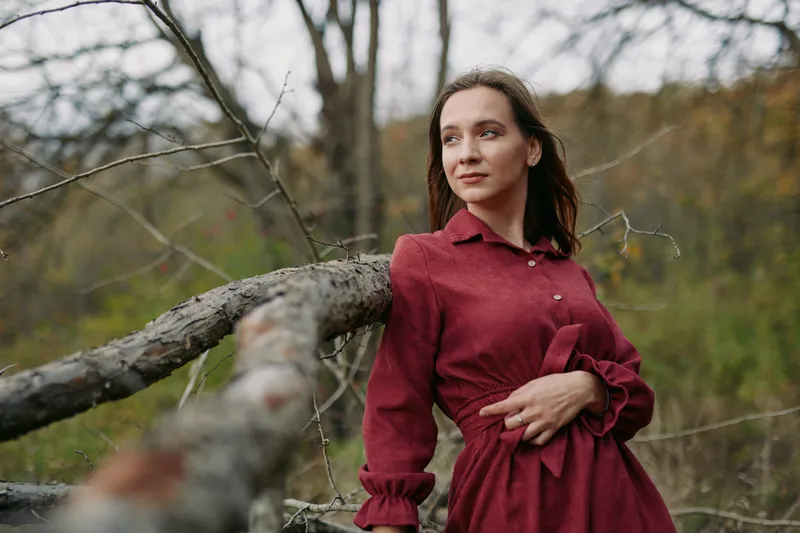A young woman in a simple burgundy dress leans on a fallen tree in an open woodland, portraying quiet authenticity and trustworthiness through candid posture and soft natural light.